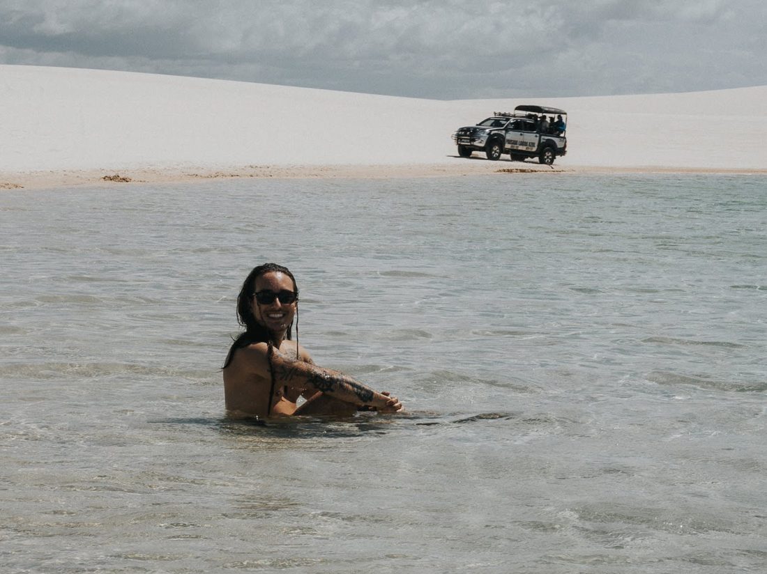 Ric sorrindo dentro de uma lagoa nos Lençóis Maranhenses, com as dunas brancas e um 4x4 ao fundo